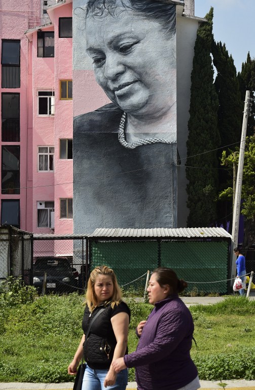 Women walk near a building decorated with a mural of Icelandic artist Guido Van Helten  at a poor neighborhood in Ecatepec, Mexico on August 25, 2016.  Dozens of murals were painted on buildings in a poor neighborhood in Ecatepec, on the route of a new cable car that will run this year. / AFP PHOTO / RONALDO SCHEMIDT / TO GO WITH AFP STORY BY JENNIFER GONZALEZ