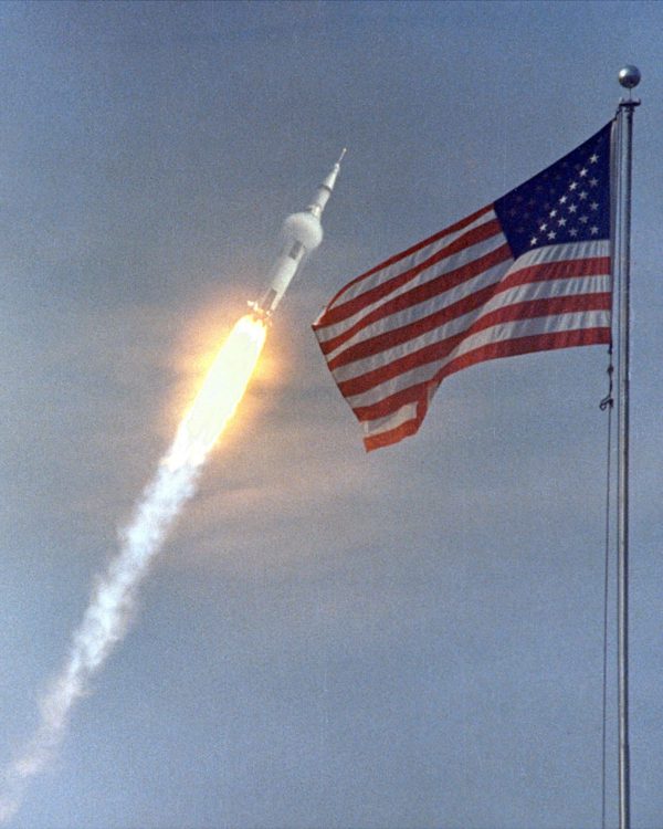 La bandera estadounidense al frente mientras en el fondo se ve el lanzamiento del Apollo 11: la primera misión de aterrizaje en la Luna en 1969.