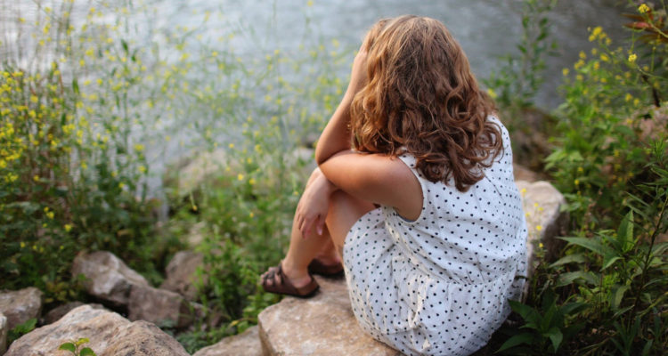 En la imagen, una mujer está sentada de espaldas apoyando su cabeza en sus manos. La chica se encuentra en un lago y usa un vestido por las altas temperaturas.