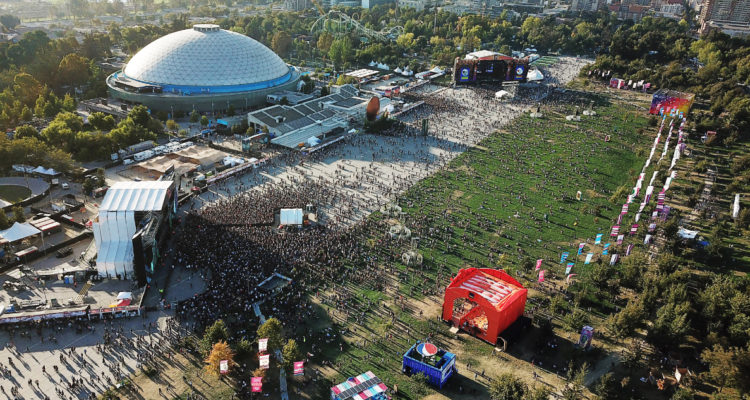 Se ve uno de los primeros días del evento en el Parque O’higgins en ediciones anteriores, la vista, desde altura, muestra como hay miles de personas en el espacio donde hay dos de los tres escenarios del evento.