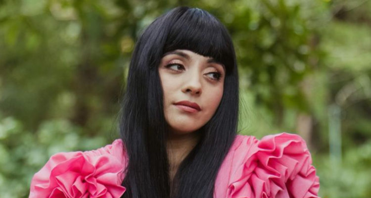 La cantante aparece con una leve sonrisa mirando hacia un costado. Está con su cabello negro y usa un vestido holgado en medio de un bosque.