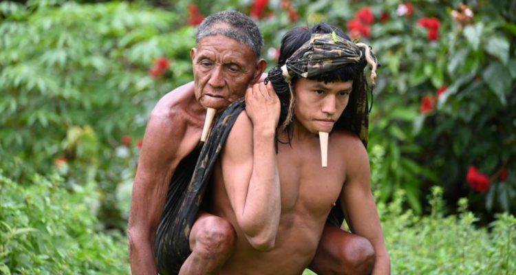 El joven Zo’é de 24 años lleva a su padre sentado en su espalda y lo sostiene con ayuda de un gran manto de tela atado a su cabeza.