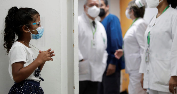 Una niña espera en un pasillo mientras que se ven a dos enfermeras y un médico al fondo.