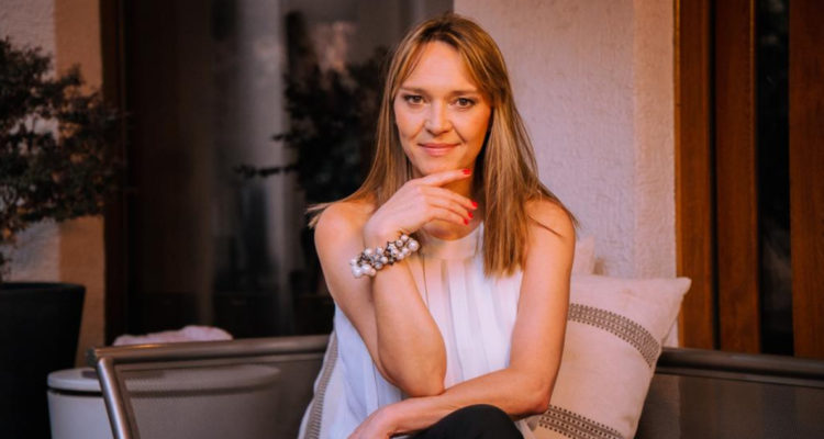 Carola Brethauer aparece sentada en un sofá con las pernas cruzadas. Apoya su rostro en su mano izquierda mientras da una sonrisa leve.