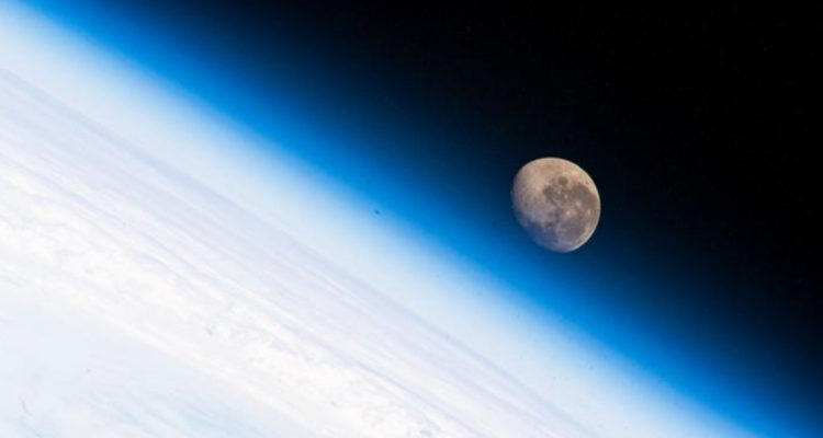 Una fotografía donde se ve parte de la atmosfera terrestra mientras que de fondo se ve la luna