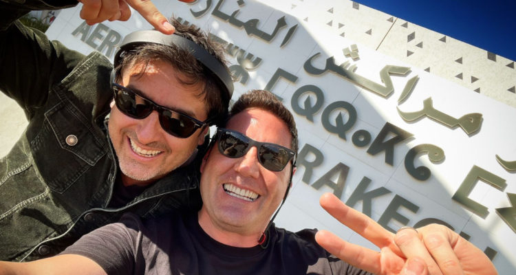 El actor Jorge Zabaleta abrazado a Pancho Saavedra en uno de sus viajes de Socios por el Mundo. Ambos usan lentes de sol, sonríen y hacenel signo de la paz con sus dedos.