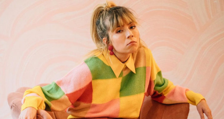 Jennette McCurdy está sentada en un sofá con un rostro serio, pese a la gran cantidad de colores alegres a su alrededor.