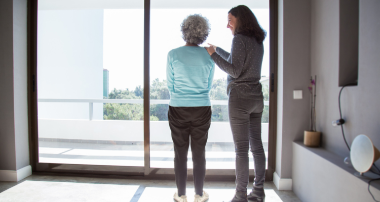 Mujer mirando por una ventana con nostalgia acompañada de su hija. Descubren factor que podría explicar por qué las mujeres desarrollan más el Alzheimer
