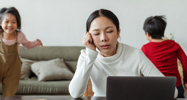 Una mujer trabaja en su notebook mientras dos de sus hijos juegan en su espalda. En la actualidad, muchas madres sufren de carga mental sin darse cuenta.