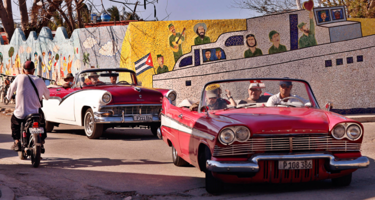 Fotografía muestra una calle de Cuba con automóviles y muros pintados con imágenes de la Revolución Cubana.