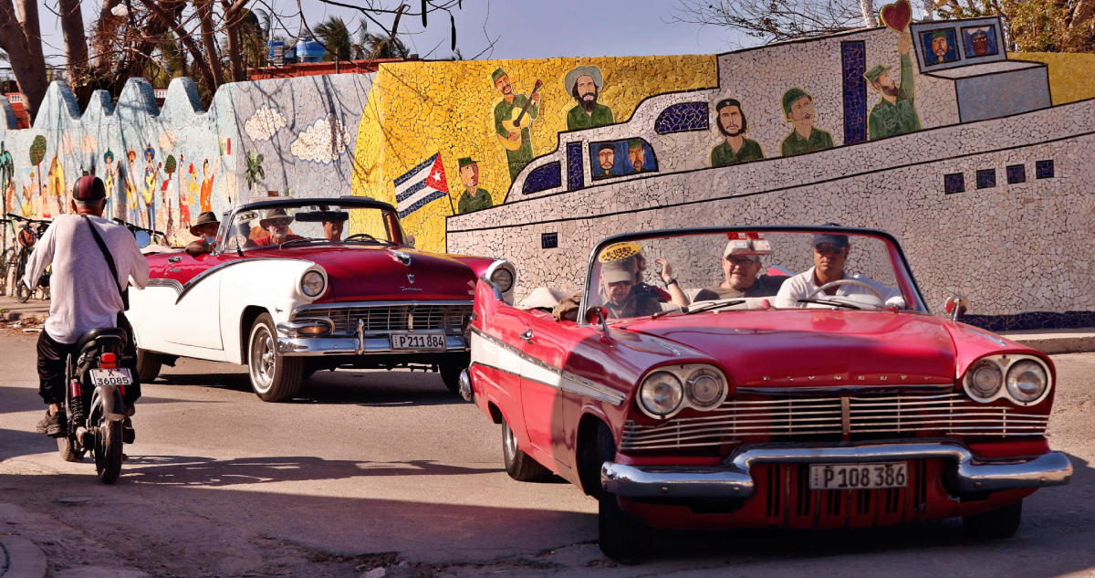Fotografía muestra una calle de Cuba con automóviles y muros pintados con imágenes de la Revolución Cubana.