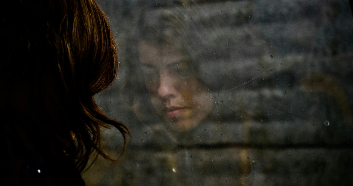 Mujer triste mirando por una ventana, en la que se ve su reflejo.