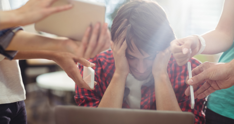 Joven frente a un computador, tomándose la cabeza, mientras es rodeado de personas con celulares y tablets.