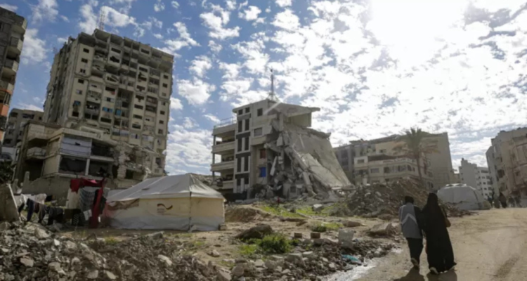 Personas caminando frente a carpa e inmuebles destruidos en Gaza.