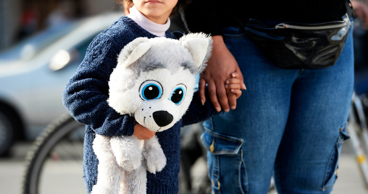 Foto de contexto de niña con un peluche de la mano de una persona adulta.