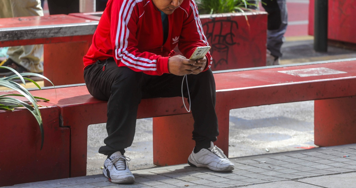 Foto de contexto de desempleo. Persona sentada en una banca mirando su celular.